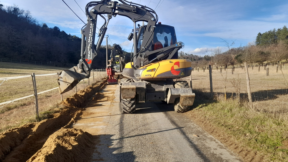 Une pelle à pneus ouvre une tranchée en bord de route, avec des matériaux excavés sur le côté.